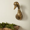 Bronze duck head sculpture on a wooden surface with greenery against a plain wall.