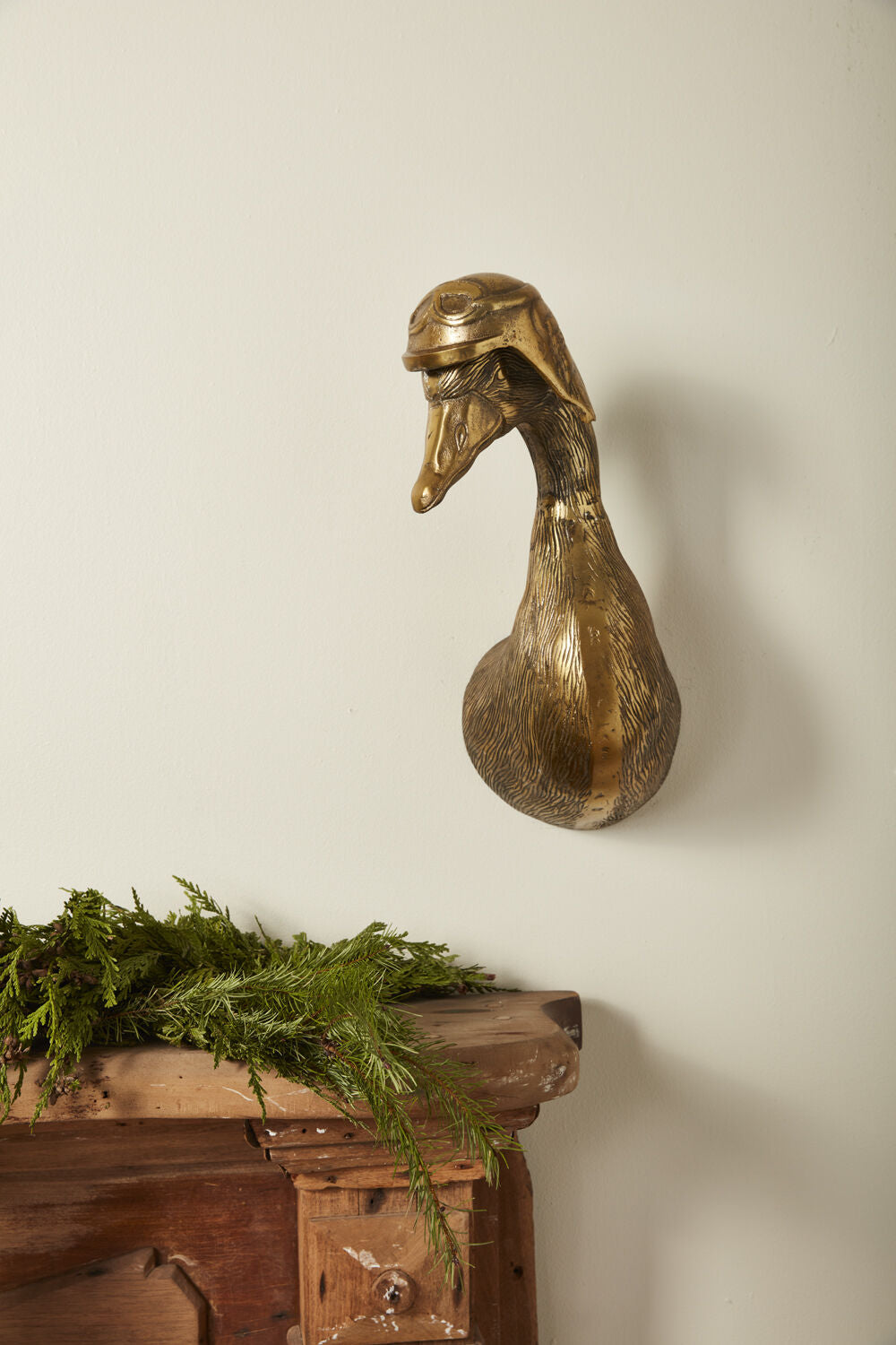 Bronze duck head sculpture on a wooden surface with greenery against a plain wall.